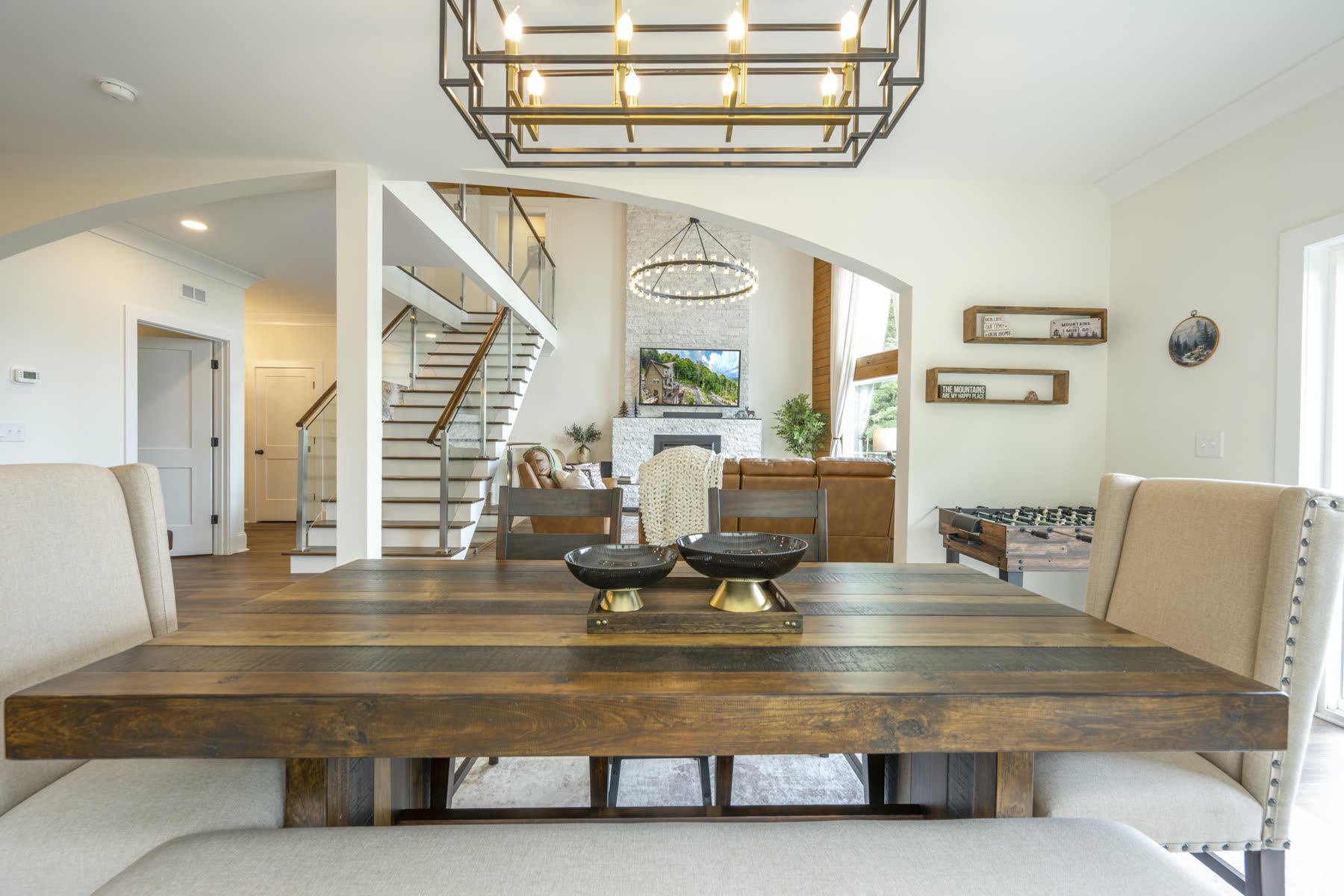 Rustic wood dining table for eight beneath a black and brass chandelier, with a foosball table and staircase in the background